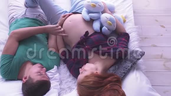 有趣的怀孕夫妇相爱地躺在床上快乐的父亲爱抚妻子的肚子生育观念怀孕视频的预览图