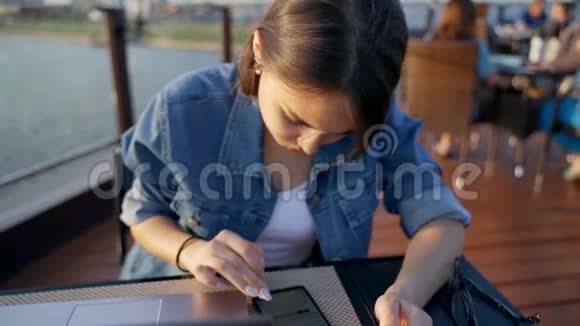 这个女孩正在智能手机上打字一个穿着牛仔夹克的黑发女人坐在一张桌子旁手提电脑和一部智能手机视频的预览图