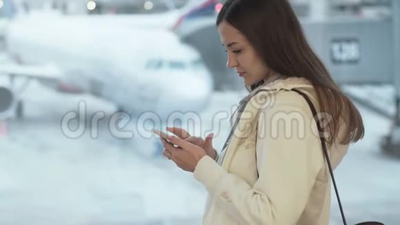 侧视美丽的女孩留着长发站在机场的窗口在飞机的背景上发短信视频的预览图