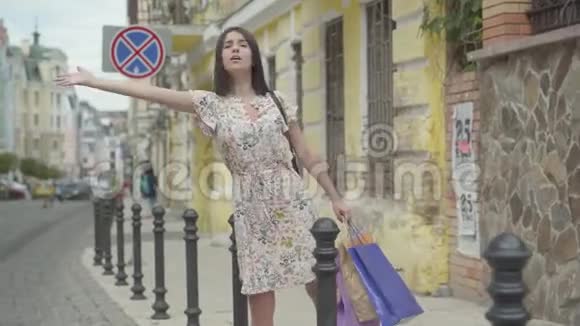 穿着夏装手里拿着购物袋想在街上搭出租车的漂亮年轻女子无忧无虑视频的预览图