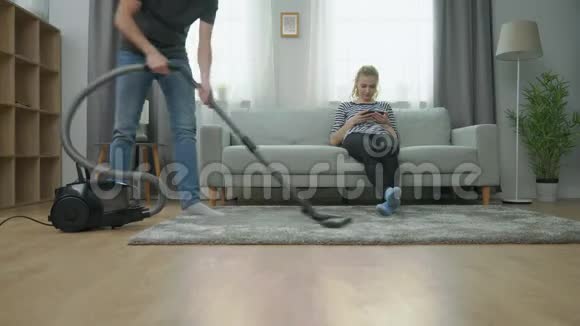 男人在地板上吸尘他的女朋友在沙发上用智能手机视频的预览图