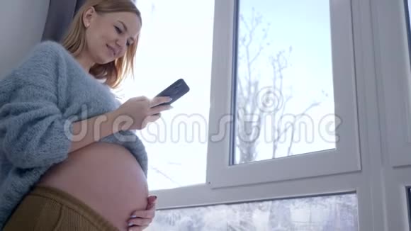 准女孩在家中站在大窗户旁用手机抚摸肚子视频的预览图