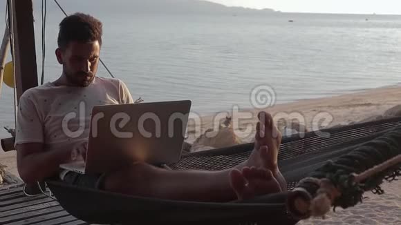 一个英俊的年轻人在海滩上的吊床上用笔记本电脑工作视频的预览图