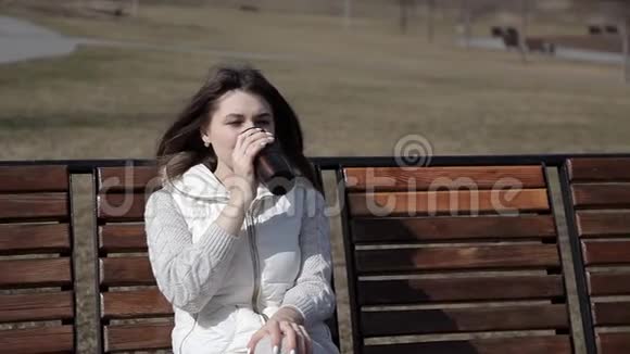 一个漂亮的女孩坐在长凳上喝着公园里的热杯咖啡烧着嘴唇视频的预览图
