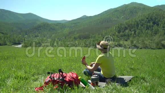 那个拿着旅游棒和一个红色背包的旅行者的女人坐在草地上看着田里的玻璃视频的预览图