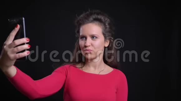 白种人女人在黑色背景下微笑着用智能手机制作自拍照片的肖像视频的预览图