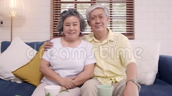 在家里客厅的沙发上放松时亚洲老年夫妇感到快乐的微笑和看着相机视频的预览图