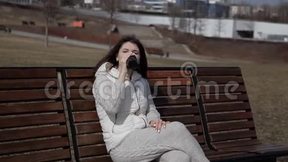 公园里一个漂亮的女孩坐在长凳上喝着热杯子里的咖啡打电话给别人视频的预览图
