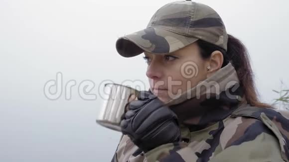 一个身穿迷彩服戴着帽子的漂亮姑娘在一片雾蒙蒙的河岸上喝着铁杯里的热茶视频的预览图