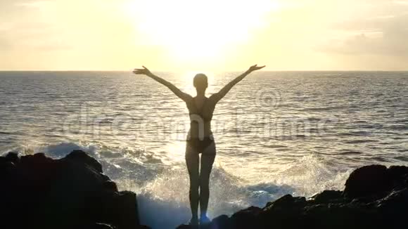 日落时分一位妇女举起手臂在空中观察海浪撞击岩石和喷洒视频的预览图