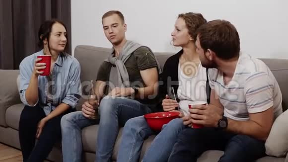 开朗的年轻男女朋友带着饮料正坐在家里的沙发上吃着红碗里的爆米花年轻人视频的预览图