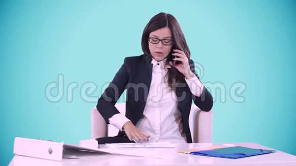 蓝色背景的年轻黑发女人坐在办公室的办公桌后面在打电话桌子上有文件视频的预览图
