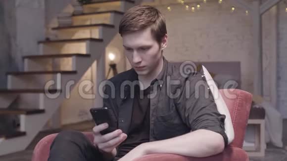 年轻男子使用智能手机坐在家里的双人公寓扶手椅上身后有花环股票视频的预览图