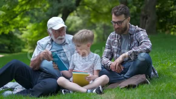 三代幸福的家庭父亲祖父和金发儿子坐在公园的草地上带着书学读书视频的预览图