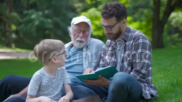 三代幸福的家庭父亲祖父和金发儿子坐在公园的草地上带着书学读书视频的预览图
