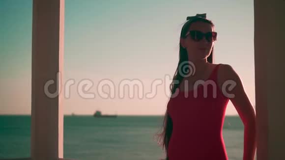 一个穿着红色的一件泳衣和眼镜的怀孕女孩站在海滩上穿着白色的混凝土凉亭视频的预览图