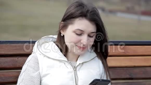 公园里的漂亮女孩坐在长凳上喝着杯子里的咖啡打着电话透过眼睛看着视频的预览图