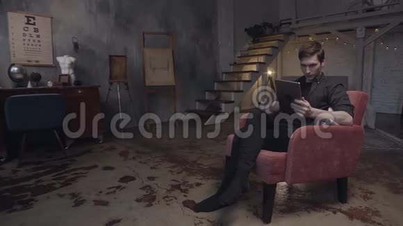 英俊的年轻人坐在红色扶手椅上使用数字平板电脑库存录像一个人在看他的装置视频的预览图