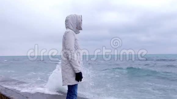 穿着白色夹克的女人站在海滩上的海滨看着暴风雨的波浪侧视视频的预览图