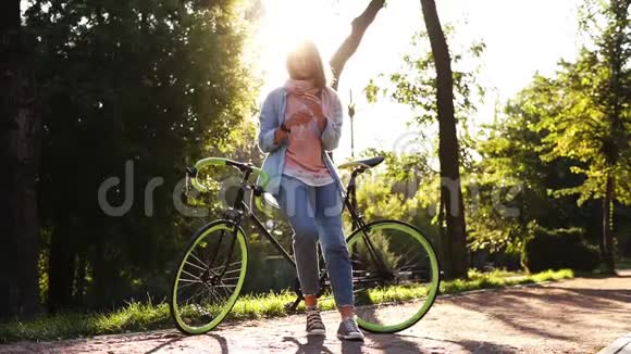 可爱的女孩从户外的瓶子里喝着清澈的水靠在她徒步旅行的绿色车轮自行车上美丽美丽视频的预览图