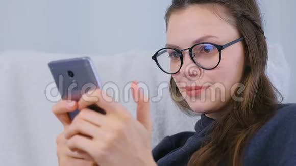 戴着眼镜的年轻黑发女人坐在扶手椅上用手机聊天视频的预览图