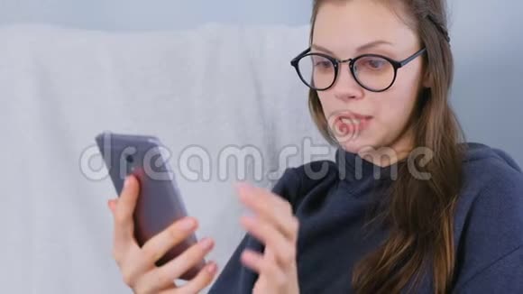 戴着眼镜的年轻黑发女人坐在扶手椅上用手机聊天视频的预览图