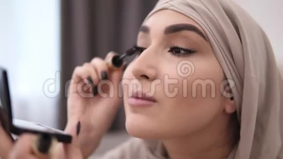 漂亮的穆斯林女性专业化妆用睫毛膏刷睫毛戴着米色头巾白墙开启视频的预览图
