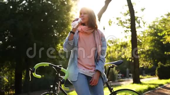 漂亮的女孩从户外的瓶子里喝着清澈的水靠在她徒步旅行的绿色车轮自行车上美丽美丽视频的预览图