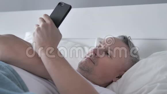 白发男子躺在床上用智能手机视频的预览图
