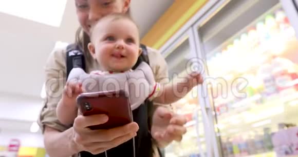 吊带婴儿的女士在商店里使用智能手机视频的预览图