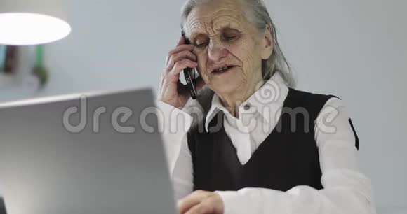 一位长着深深皱纹的老妇人坐在笔记本电脑旁用手机说话视频的预览图