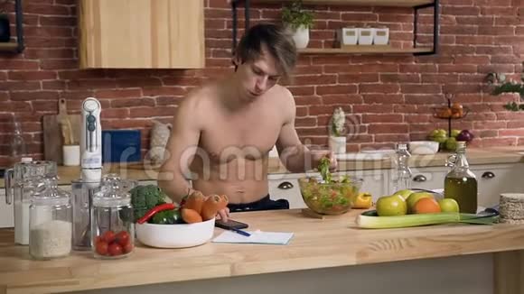 身材赤裸的年轻人坐在厨房的桌子后面一边吃纯素沙拉一边用智能手机聊天视频的预览图