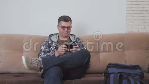 一个中年人坐在酒店房间或公寓的沙发上用智能手机上网旁边是一个视频的预览图