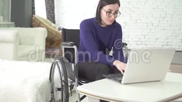 带眼镜的年轻残疾妇女坐在轮椅上手提电脑在家里面带微笑视频的预览图