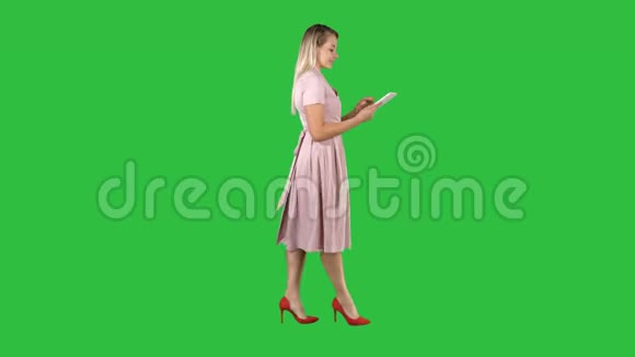 金发碧眼的粉红色连衣裙行走和使用平板上的绿色屏幕铬键视频的预览图
