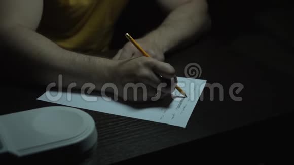 一个穿黄色T恤的人在黑暗中的桌子上工作一个人用铅笔在纸上用手写字特写镜头视频的预览图