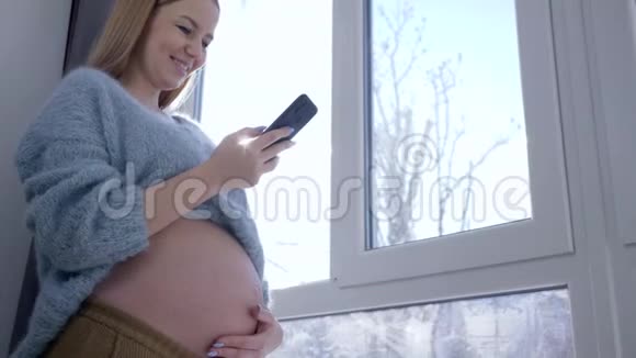 现代怀孕大肚子的漂亮孕妇在冬天阳光下对着窗户看智能手机视频的预览图