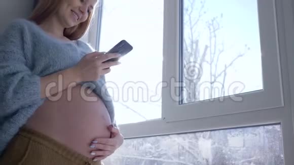 微笑的大肚子孕妇在阳光下对着窗户等宝宝的时候使用了现代移动技术视频的预览图