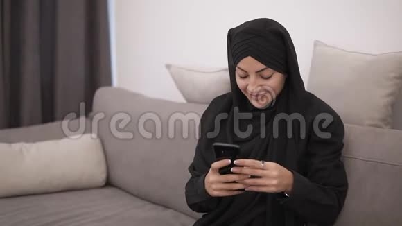 美丽的穆斯林妇女戴着黑色头巾坐在沙发上看着她的智能手机微笑着现代lof视频的预览图