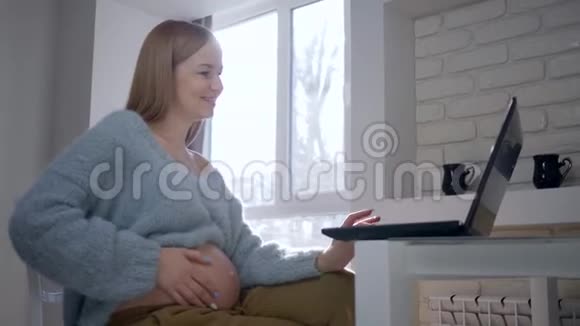 视频连接怀孕的女性使用笔记本电脑与朋友在互联网上聊天并显示赤裸的腹部坐着视频的预览图