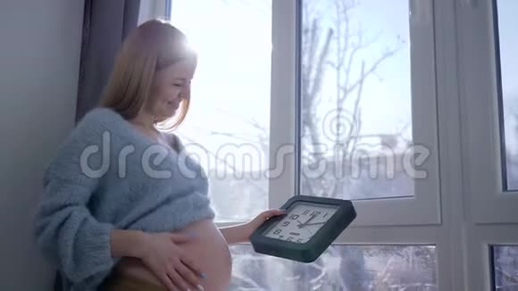 大肚子的年轻准妈妈在冬日的阳光下手拿着挂钟对着窗户视频的预览图