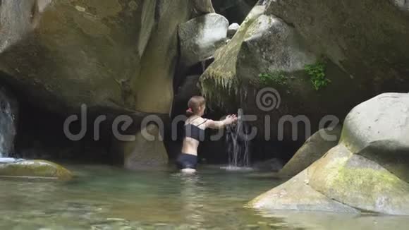 美丽的女孩把双手放在山湖瀑布下在绿色的热带森林里上面有巨石和苔藓视频的预览图