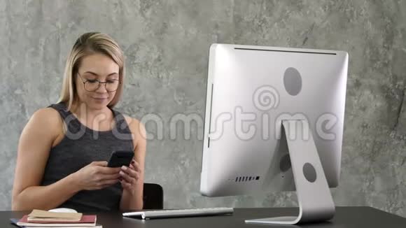 年轻的微笑商务女性在办公室的电脑附近使用智能手机视频的预览图