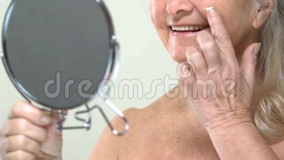 老妇人在小镜子前涂抹抗衰老面霜身体护理视频的预览图
