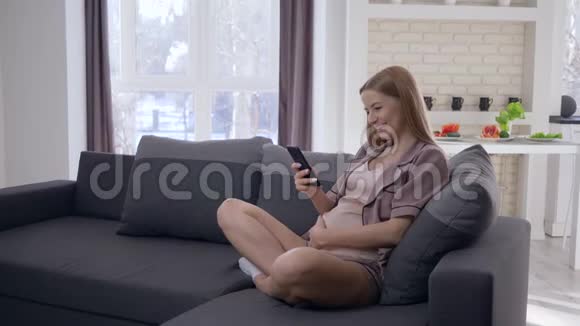 现代怀孕微笑怀孕的女性使用手机和抚摸肚子未来的婴儿坐在家里的沙发上视频的预览图