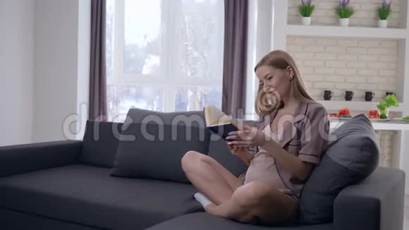 年轻漂亮的孕妇大肚子读着有趣的书抚摸着肚子未来的孩子坐在沙发上视频的预览图