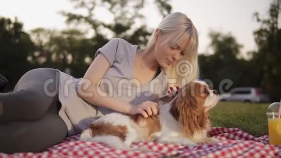 一个穿着灰色紧身裤和亮灰色T恤的金发女人和她的小狗躺在格子地上的肖像爱抚和抚摸视频的预览图