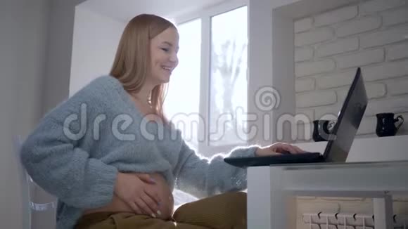 怀孕快乐怀着光着肚子微笑的准女孩用坐在笔记本电脑前的智能电脑视频的预览图