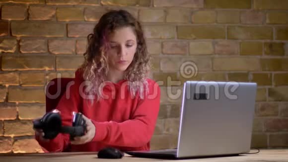 穿红色连帽衫的年轻女孩的肖像戴上耳机在Briken墙背景上观看笔记本电脑视频的预览图
