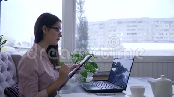 戴眼镜的女商人用手提电脑在桌旁的笔记本上用蓝色记号笔写笔记视频的预览图